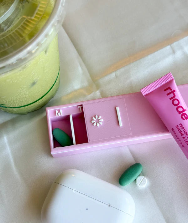Pink pill organizer with green pills, white device, and jar on a light surface