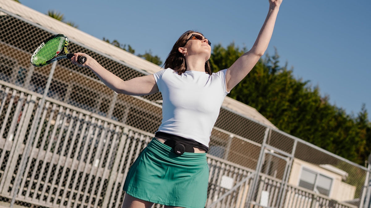 A woman on an outdoor tennis court serves a tennis ball, wearing a white polo shirt, a green skirt, and white sneakers under a clear blue sky. Relieving her pain with Tap 2.0 TENS evidence-based device. 